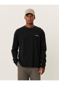 Svart langermet skjorte i bomull med rund hals. Har en liten hvit logo på brystet og er kombinert med en beige caps.