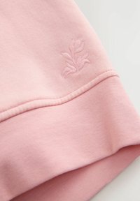 Roza majica iz tkanine z gladko teksturo. Ima subtilno vezeno cvetlično obliko v spodnjem levem kotu.