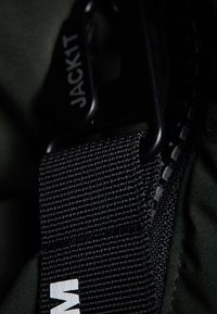 Černý tkaný popruh s texturovaným povrchem, připevněný k tmavě zelené tkanině. Obsahuje černou plastovou sponu s logem "JACK1T".