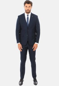 Completo blu con una vestibilità sartoriale, caratterizzato da rever a punta, due bottoni e tasche frontali. Indossato con una camicia bianca e una cravatta scura, completato da scarpe nere lucide.