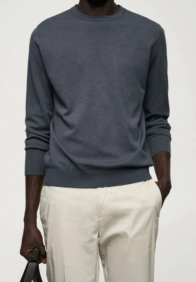 Homme portant une chemise à manches longues gris foncé et un pantalon beige clair, tenant un sac de couleur sombre dans la main gauche.