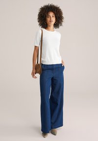 Kvinna med lockigt hår som bär en vit texturerad kortärmad topp, vida blå jeans, beige skor, brun axelväska och guldörhängen.