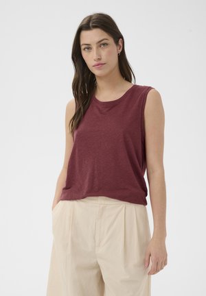 Femme aux longs cheveux bruns portant un haut bordeaux sans manches et un pantalon beige plissé à jambes larges, debout devant un fond blanc uni.