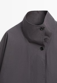 Ciemnoszary płaszcz z wysokim kołnierzem, z dwoma widocznymi czarnymi guzikami i wyraźnymi przeszyciami strukturalnymi na przedniej części ramion.