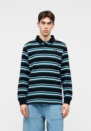 Νεαρός άνδρας που φοράει μακρυμάνικο πόλο πουκάμισο με ριγέ μαύρο, τυρκουάζ και άσπρο, με μπλε παντελόνι κοτλέ, στέκεται μπροστά σε απλό λευκό φόντο.