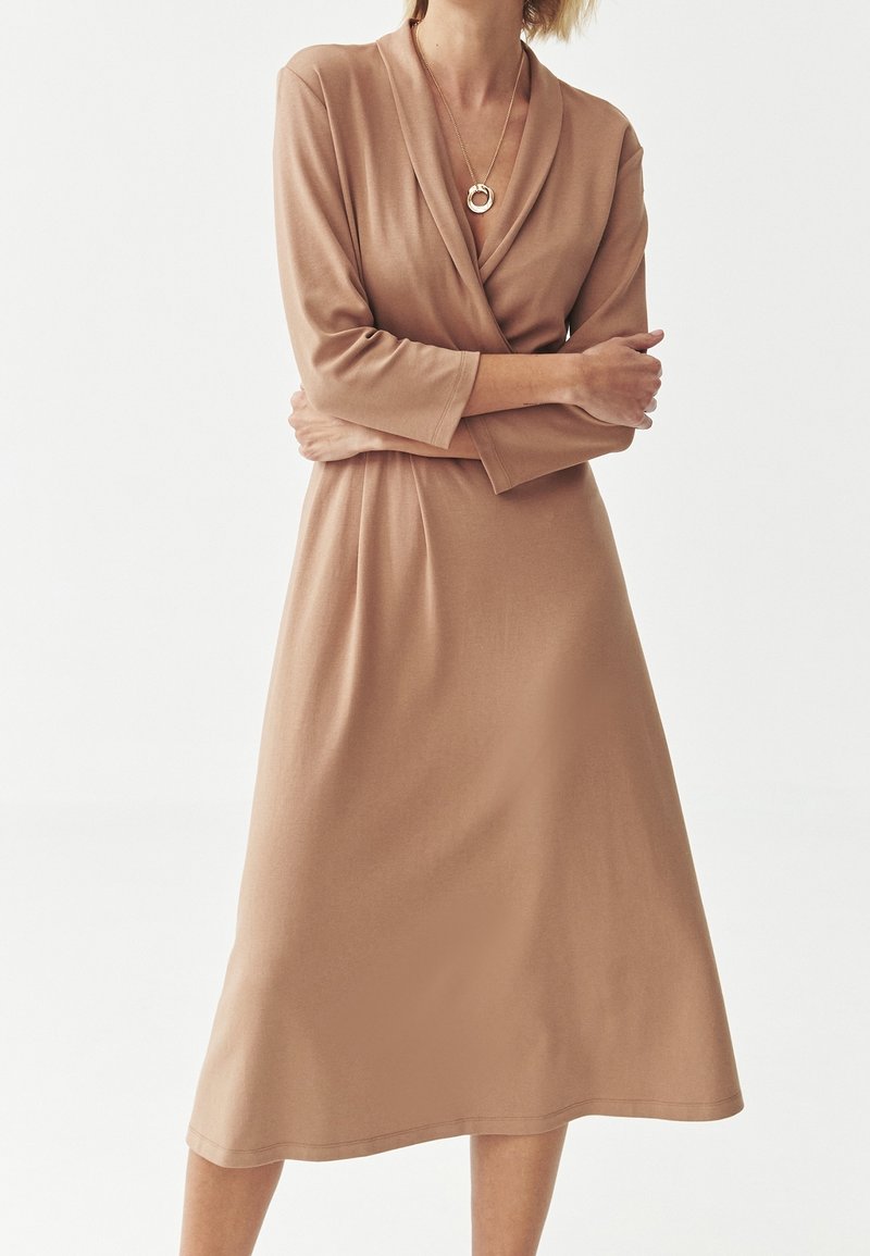 Robe à manches longues beige avec un design croisé, longueur au genou et texture douce. Présente un décolleté en V et une coupe décontractée. Accentuée par un collier circulaire doré.