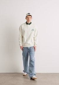 Felpa color crema con colletto verde e logo, jeans azzurri, sneakers bianche e un cappellino bianco con logo. Outfit casual.