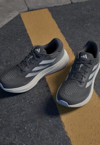 Baskets de sport grises avec une tige texturée, des accents blancs et une semelle rembourrée. Présente des motifs perforés à la surface et un système de laçage.