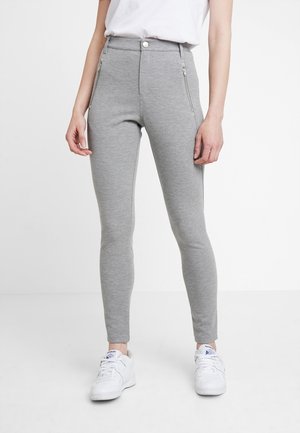 Personne portant un pantalon gris ajusté avec des fermetures éclair à l'avant et des baskets blanches, debout devant un fond blanc uni.