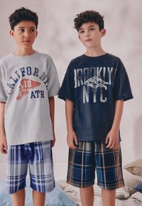 Deux garçons se tiennent côte à côte, l'un portant un t-shirt gris avec un logo ailé et un short à carreaux bleu, l'autre un t-shirt bleu foncé avec une inscription blanche et un short à carreaux.