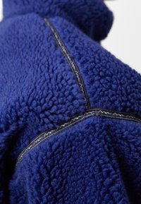 Marineblå fleecejakke med en tekstureret, fluffy overflade. Har et sort zigzag-mønster langs sømmen for ekstra detaljer.