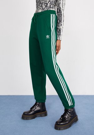 Person iført grønne Adidas joggingbukser med hvide striber ned langs siderne og sorte plateau snørestøvler.