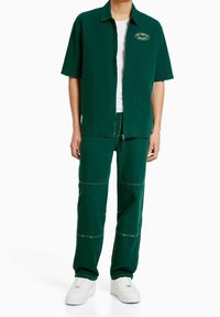 Camisa verde de manga corta con un parche bordado en el pecho; pantalones verdes a juego con costuras visibles; zapatillas blancas.