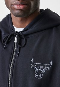 Sudadera con cremallera negra que presenta un logo de toro plateado en el pecho, capucha con cordón y material de algodón suave. Cierre con cremallera con acentos plateados.
