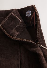 Close-up of dark brown corduroy pants waistband with button, zipper partially open, and internal elastic adjustable strap visible.