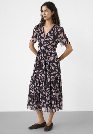 Kvinde med langt mørkt hår iført en sort midi-kjole med lyserødt bladmønster og korte ærmer, kombineret med mørkebrune flade sko.