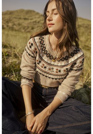 Beige villa neule, jossa on kuvioitu kaula-aukko, ruskeilla, mustilla ja oransseilla yksityiskohdilla. Denim-farkut kultaisella vyöllä, ulkotila.