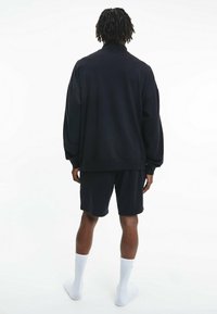 Sudadera negra con hombros caídos, puños acanalados y pantalones cortos negros a juego. Modelo con calcetines blancos hasta el tobillo, vistos desde atrás.