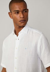 White short-sleeve shirt made of linen with a textured finish. Features a button-down collar and a small blue embroidered logo on the chest.