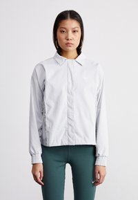 Grå Nike-jakke med krage, elastiske cuffs og slim fit. Har en liten hvit logo på venstre bryst og sidelommer.