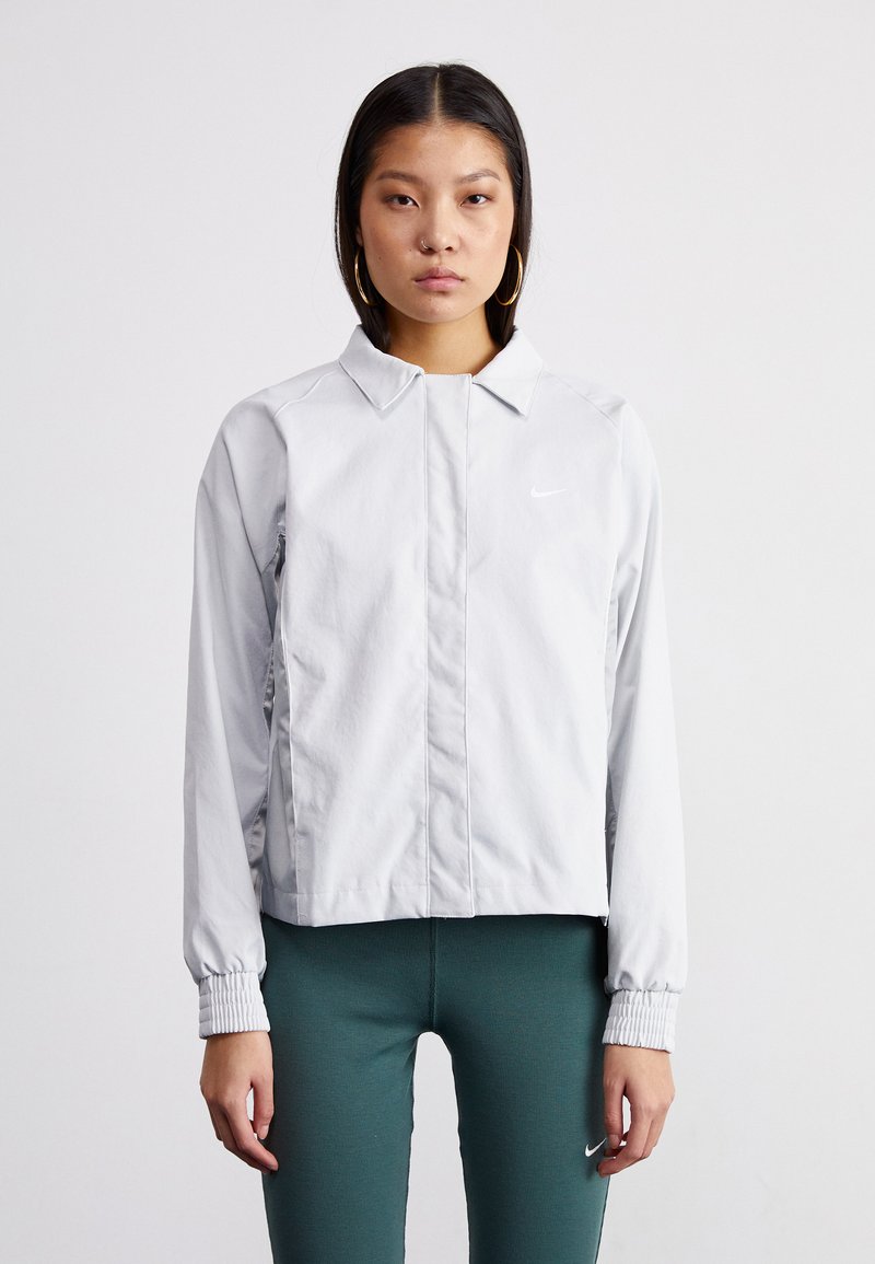 Grå Nike-jakke med krage, elastiske cuffs og slim fit. Har en liten hvit logo på venstre bryst og sidelommer.