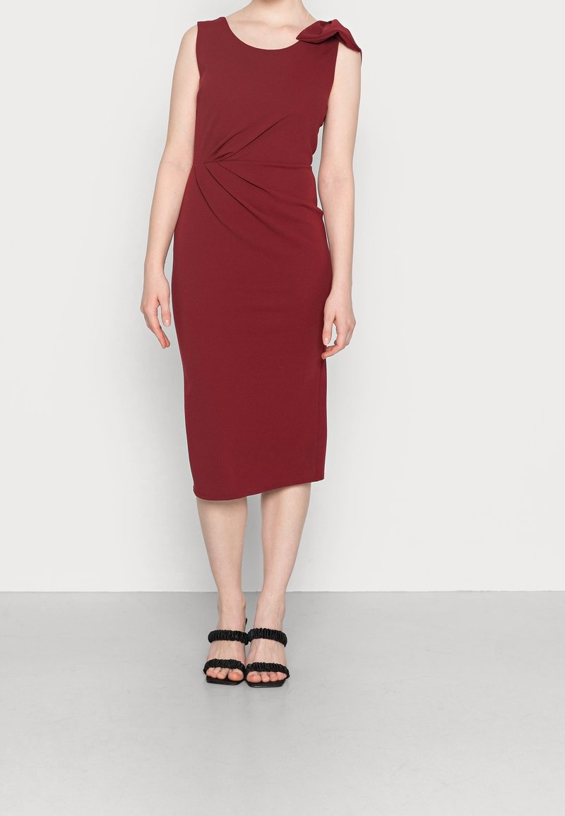 Robe ajustée bourgogne avec un détail de nœud sur le côté, sans manches et jupe au genou ; associée à des sandales noires à lanières.