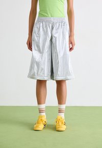 Shorts de sport argentés et brillants avec une taille élastique, coupe au genou et poches latérales, associés à des chaussures jaunes et des chaussettes rayées.
