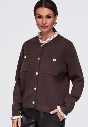 Femme portant un cardigan marron boutonné sur un chemisier blanc à volants avec des pois noirs, assorti à un pantalon en cuir noir.
