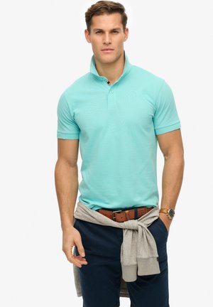 Helles türkisfarbenes Poloshirt mit Kragen, zwei Knöpfen und gestreiften Akzenten. Getragen mit marineblauen Shorts und einem grauen Sweatshirt, das um die Taille gebunden ist.