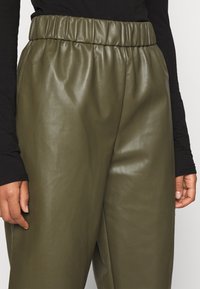 Pantaloni in ecopelle verde oliva con un bordo elastico in vita, caratterizzati da una texture liscia e una vestibilità comoda. Mano parzialmente visibile.