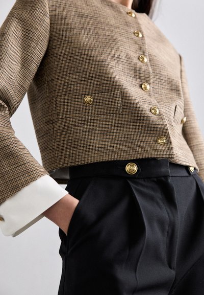 Bruin houndstooth cropjasje met gouden knopen, witte manchetdetails, gecombineerd met hoogzittende zwarte geplooide broek met een vergelijkbare knop.