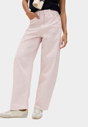 Donna che indossa pantaloni rosa chiaro a gamba larga con tasche anteriori e sneakers beige casual, top nero e sciarpa crema su una spalla.