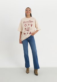Crème oversized t-shirt met rode graphic tekst en woestijn-thema illustraties, gecombineerd met blauwe flared jeans en bruine hoge hakken laarzen.