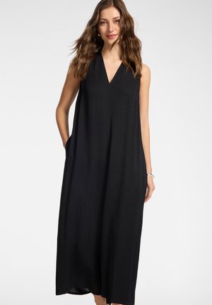 Femme aux cheveux bruns ondulés portant une robe maxi noire sans manches à col en V, les mains dans les poches latérales, souriante sur un fond blanc uni.