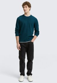 Türkis gestreifter Pullover mit Rundhalsausschnitt, schwarze Jeans und weiße Sneakers. Das Model steht mit den Händen in den Taschen, leicht zur Seite gedreht, lässig gestylt.