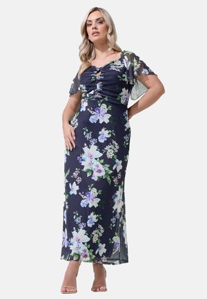 Kvinde iført en marineblå blomsterprint maxi-kjole med rynket krop og flagrende ærmer, stående med den ene hånd på hoften og neutrale hæle.