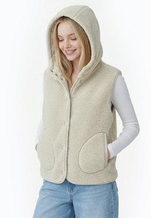Kvinde iført lysebeige sherpa-fleecevest med hætte og trykknapper, hvid langærmet skjorte og blå jeans, hænderne i lommerne, smilende ned.