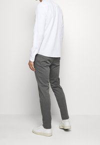 Muž stojící v šedých slim-fit kalhotách, bílé dlouhé košili a bílých teniskách před jednobarevným světlým pozadím.