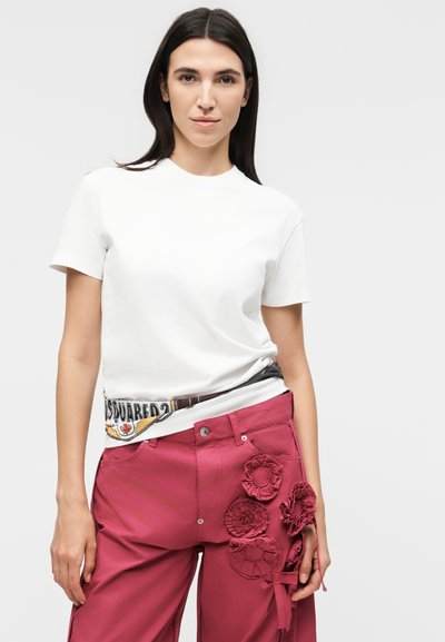 Γυναίκα με μακριά σκουρόχρωμα μαλλιά, που φοράει λευκό κοντομάνικο πουκάμισο και ροζ παντελόνια με 3D ανθισμένες εφαρμογές στο δεξί μηρό, στέκεται μπροστά σε γκρι φόντο.