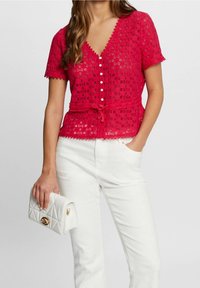 Blouse en dentelle rouge avec des manches courtes, encolure en V, bords festonnés, boutons sur le devant et taille nouée. Associée à un pantalon blanc et un sac à main matelassé.