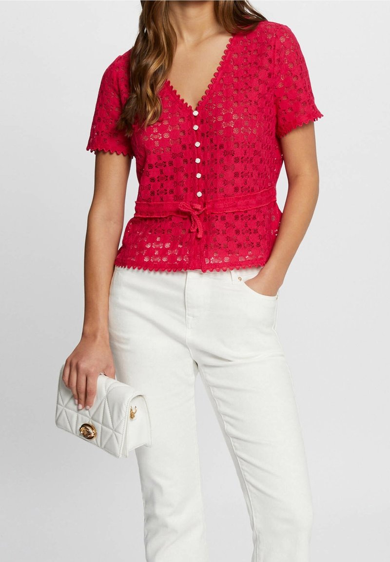 Blouse en dentelle rouge avec des manches courtes, encolure en V, bords festonnés, boutons sur le devant et taille nouée. Associée à un pantalon blanc et un sac à main matelassé.