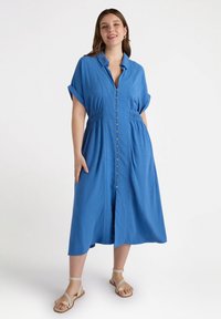 Abito azzurro cielo con bottoni, maniche corte arrotolate, vita elastica e orlo a ginocchio. Sandali con cinturini incrociati completano il look.