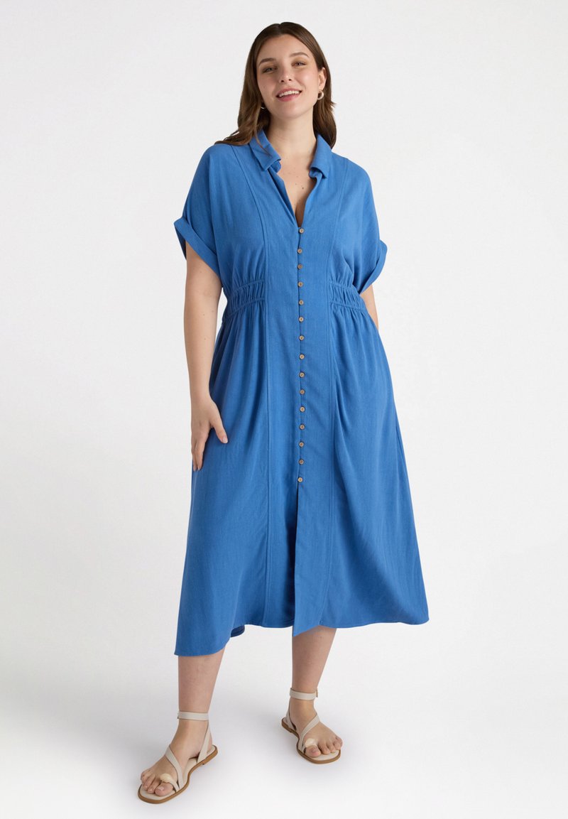 Abito azzurro cielo con bottoni, maniche corte arrotolate, vita elastica e orlo a ginocchio. Sandali con cinturini incrociati completano il look.