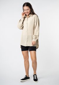Beige cotton button-up shirt with a textured finish, long sleeves, and slightly curved hem, paired with black bike shorts and black sneakers.