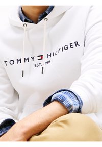 Sweat à capuche blanc avec cordons, arborant le logo "TOMMY HILFIGER EST. 1985". Surmonté de poignets de chemise à carreaux bleus. Coupe décontractée.