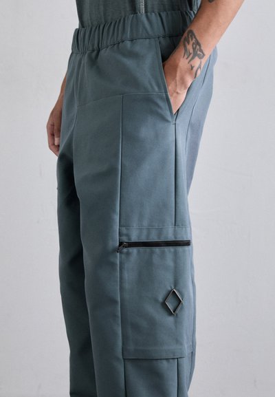 Pantalones cargo en un tono teal apagado, hechos de una tela suave. Las características incluyen una cinturilla elástica, un bolsillo con cremallera y un acento en forma de diamante.
