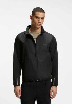 Junger Mann trägt eine schwarze Reißverschlussjacke mit BOSS-Logo, darunter ein beige T-Shirt und schwarze Hose vor einem schlichten, hellen Hintergrund.