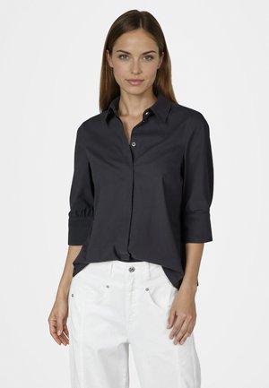 Frau mit glattem braunem Haar, die ein schwarzes, geknöpftes Hemd mit Dreiviertelärmeln und weiße Hosen trägt, steht vor einem schlichten Hintergrund.