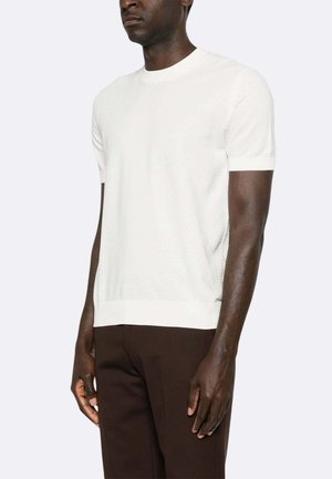 Uomo che indossa una maglia a maniche corte bianca semplice e pantaloni marrone scuro, in piedi davanti a uno sfondo chiaro uniforme.