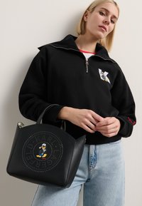 Bolsa preta feita de pele com um design circular decorado com rebites e um motivo central de personagem. A bolsa tem bordas arredondadas e alças curtas.
